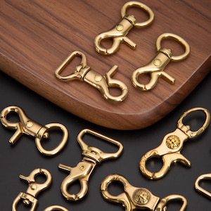 May include: Several brass-colored swivel snap hooks are arranged on a wooden surface. The hooks have a spring-loaded clasp and a circular ring. Some hooks have decorative floral designs.