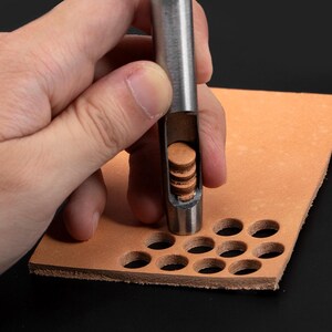 May include: A close-up of a leather punch tool with a stack of leather circles inside. The tool is being used to punch holes in a piece of brown leather.