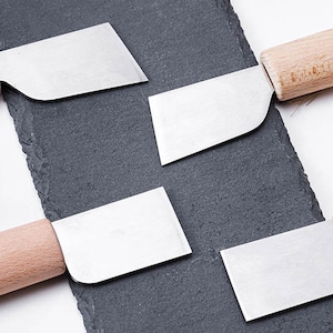 May include: Four small, rectangular, silver-colored blades with wooden handles. The blades are arranged on a black slate surface.