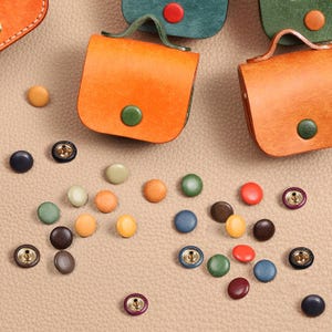 May include: Two small orange leather pouches with green snap closures and a collection of assorted colorful metal snap buttons in shades of red, green, blue, yellow, brown, and black scattered across a textured beige leather surface.