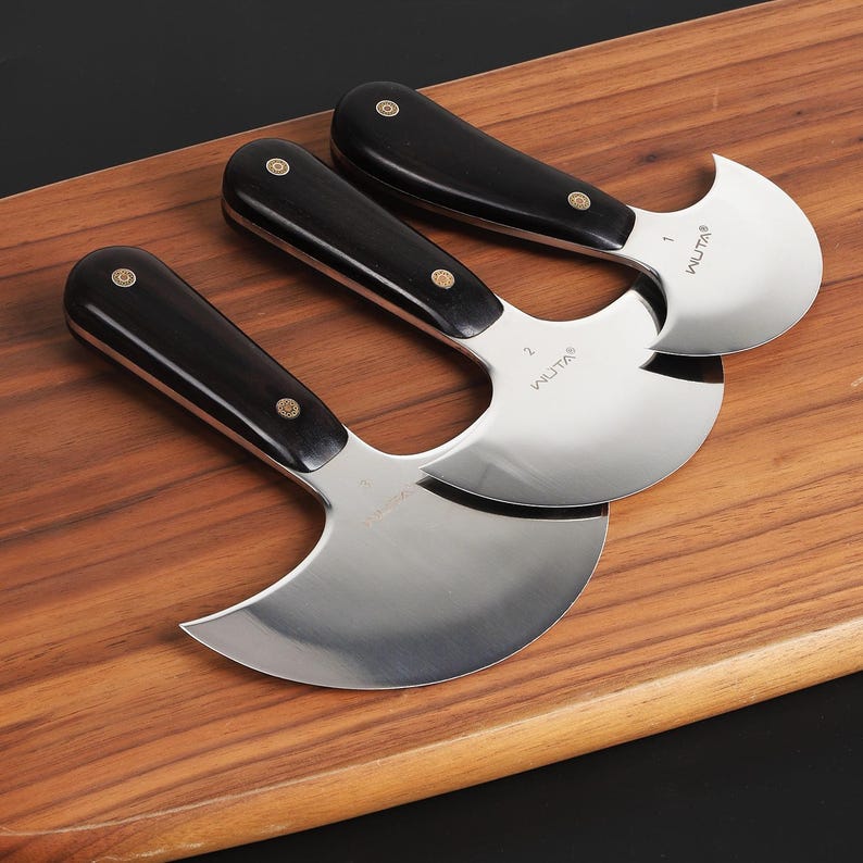 May include: Three stainless steel mezzaluna knives with black wooden handles and brass rivets are arranged on a wooden cutting board. The blades are curved and marked with the brand name "WUTA" and numbers.