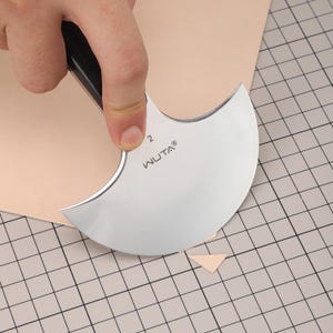 May include: A silver WUTA leather cutting knife with a black handle is used to cut a piece of tan leather. The knife has a curved blade and the number 2 is visible. The leather is on a grid cutting mat.