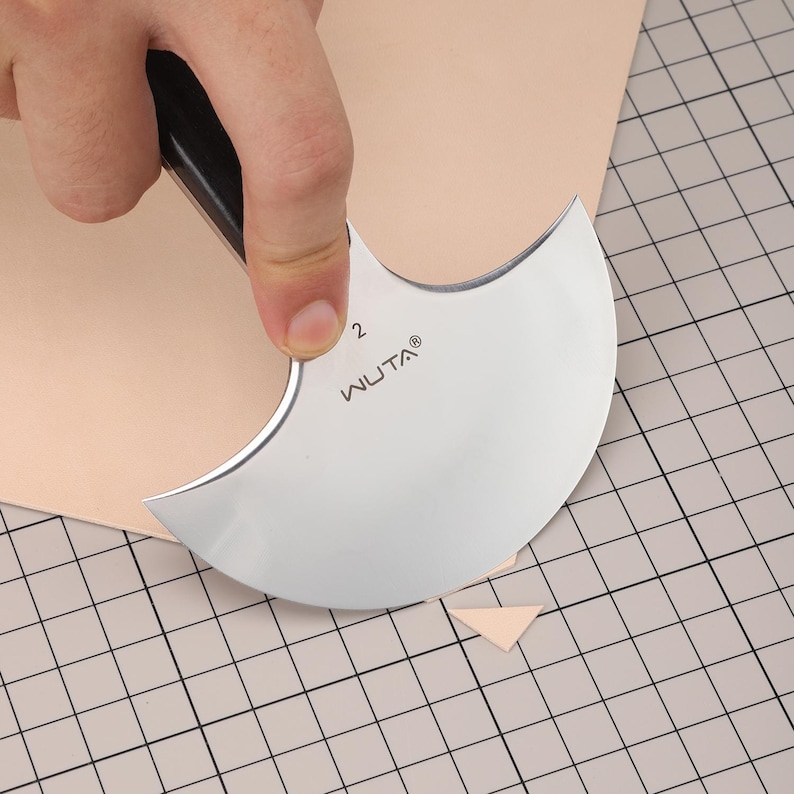 May include: A silver WUTA leather cutting knife with a black handle is used to cut a piece of tan leather. The knife has a curved blade and the number 2 is visible. The leather is on a grid cutting mat.