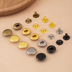 May include: A collection of metal snap fasteners in various colors, including gold, silver, and black. The fasteners are in different shapes and sizes, with both flat and domed designs. Some have a central hole for attachment.