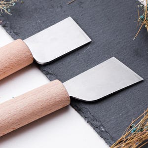 May include: Two wooden handled craft knives with metal blades. The knives are on a black slate surface.