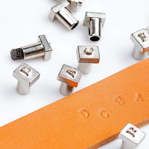 May include: A set of metal letter stamps for leather crafting. The stamps are square with a raised letter on each. The letters are A, B, C, D, and E. The stamps are shown on a piece of brown leather with the letters stamped into the leather.