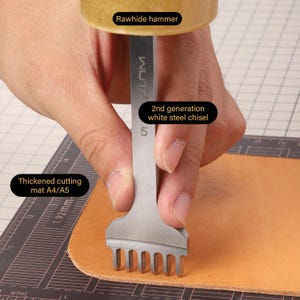 May include: A hand holds a 5-prong leather stitching chisel with a rawhide hammer above it. The chisel is labeled "2nd generation white steel chisel" and the hammer is labeled "Rawhide hammer". The chisel is being used to cut a piece of leather on a cutting mat labeled "Thickened cutting mat A4/A5".