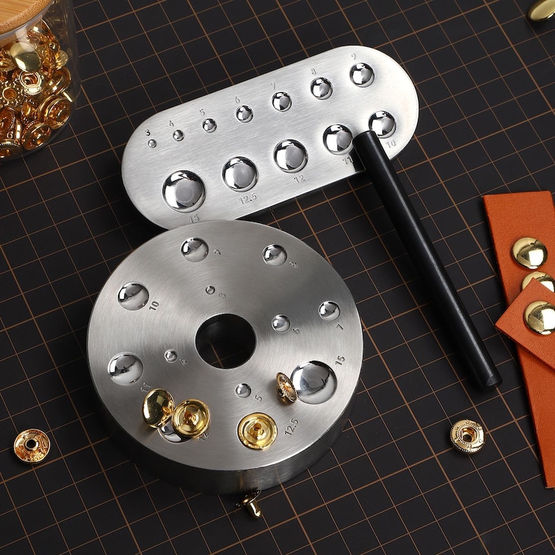 May include: Two metal tools for setting snaps, one round and one rectangular, with numbers indicating different sizes. The tools are on a black grid surface with gold snaps scattered around.