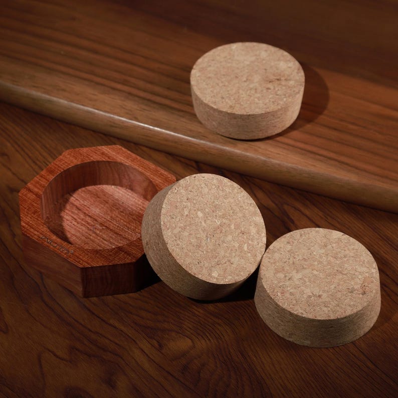 Puede incluir: Un cuenco octogonal de madera con interior marr&oacute;n rojizo se encuentra sobre una superficie de madera oscura, acompa&ntilde;ado de tres posavasos cil&iacute;ndricos de corcho. Un posavasos descansa sobre una repisa de madera, mientras que los otros dos est&aacute;n colocados cerca.