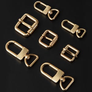 May include: A collection of gold-toned metal hardware components including three swivel lobster claw clasps with D-ring attachments and three rectangular pin buckles of varying sizes, all featuring a smooth, polished, and reflective metallic finish.
