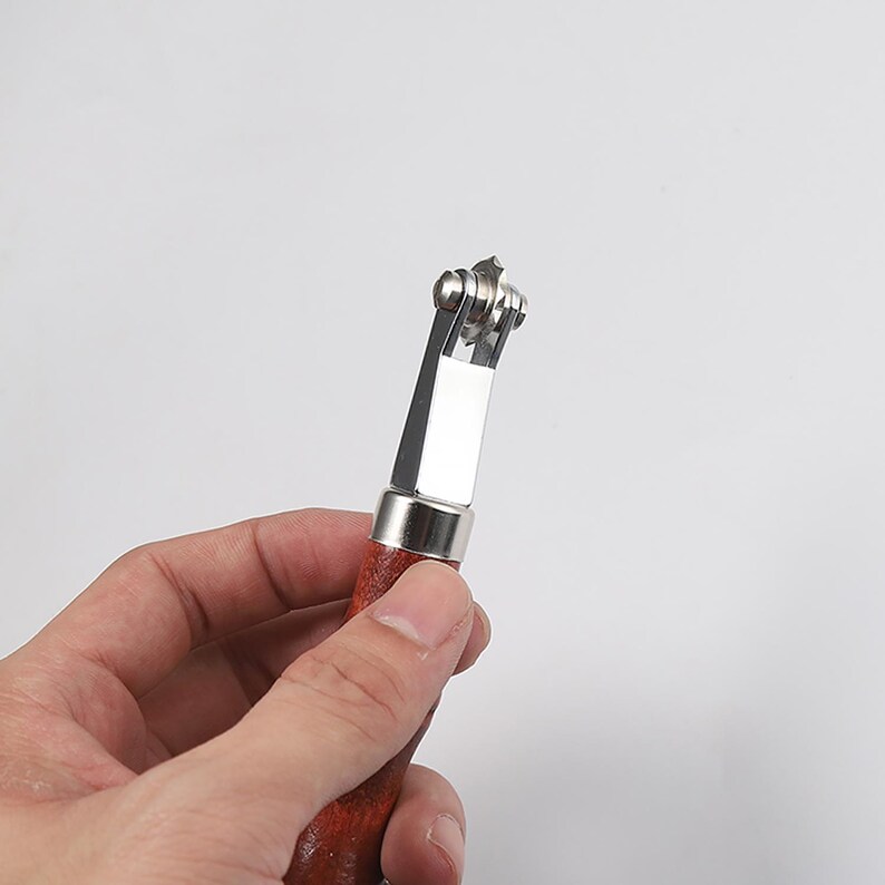 May include: A close-up of a leather working tool with a wooden handle and a metal wheel with multiple blades. The tool is held in a hand.
