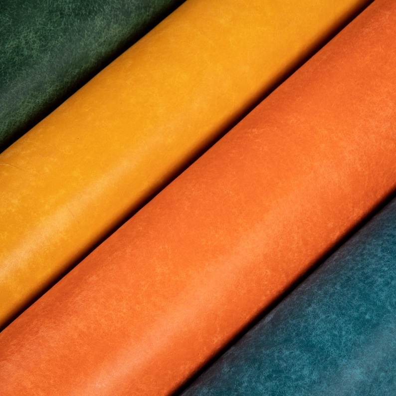 May include: Close-up of four rolls of leather in diagonal arrangement. The colors are deep green, golden yellow, bright orange, and teal blue. The leather has a textured, distressed appearance.