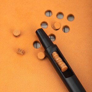 May include: A black leather crafting tool with a hollow center filled with brown leather pieces. The tool is lying on a brown leather surface with several brown leather pieces scattered around it.