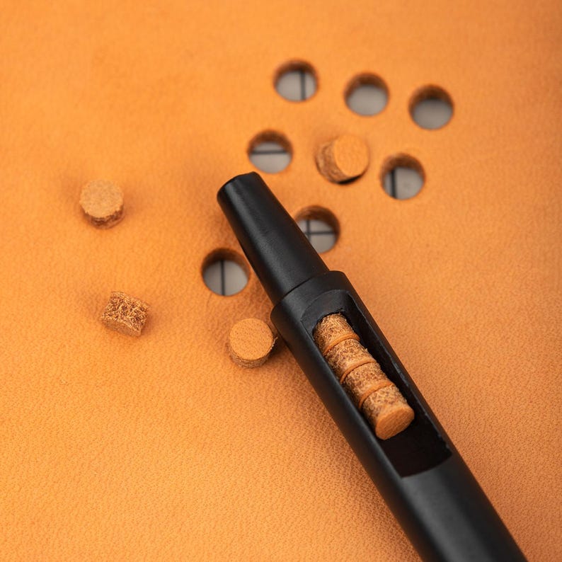 May include: A black leather crafting tool with a hollow center filled with brown leather pieces. The tool is lying on a brown leather surface with several brown leather pieces scattered around it.