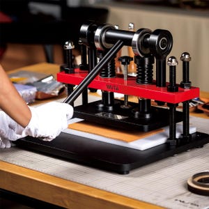 May include: A red and black leather craft tool with a lever and a metal plate. The tool is labeled "WUTA" and is being used to press a piece of leather.