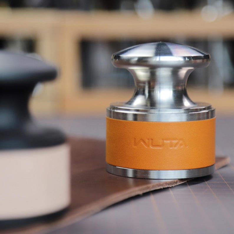 May include: A stainless steel tamper with an orange leather band embossed with "WUTA". The tamper sits on a brown leather surface. Another black tamper is slightly out of focus in the background.