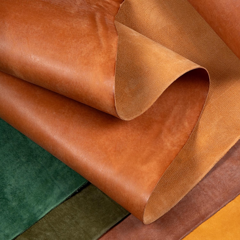 May include: Close-up of several sheets of tanned leather in shades of brown, with a hint of green and other colors. The leather has a textured surface and is folded over itself, revealing the underside.