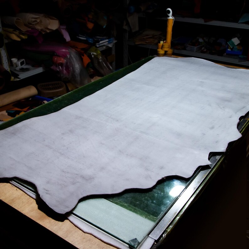 May include: A large, pale grey leather hide rests on a glass-topped table. The hide has a natural, uneven edge and a slightly textured surface. The workshop setting suggests a focus on leathercraft or related artisan work.