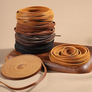 May include: A collection of flat leather cords in various shades of brown and black. One stack is arranged vertically, while two other coils are laid flat in circular patterns. The cords are smooth, flexible, and uniform in width.