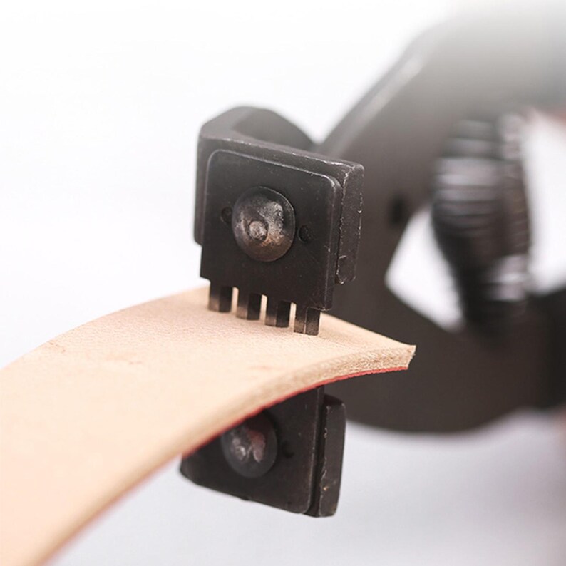 May include: Close-up of a leathercraft tool punching holes in a strip of light brown leather. The tool is black and has a series of small, evenly spaced prongs. The leather strip is curved, showing a red edge.