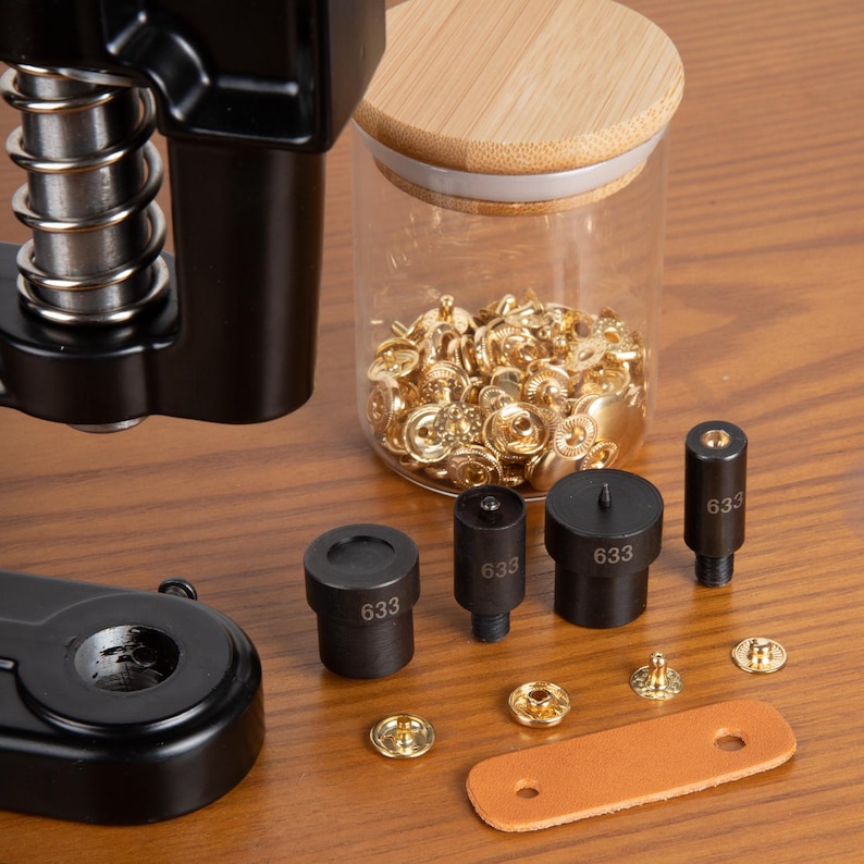 Op de afbeelding: Een zwarte drukknoop persmachine met diverse matrijzen met het label "633", een glazen pot gevuld met gouden drukknopen en een leren strip. De gereedschappen zijn gerangschikt op een houten oppervlak, klaar voor gebruik.