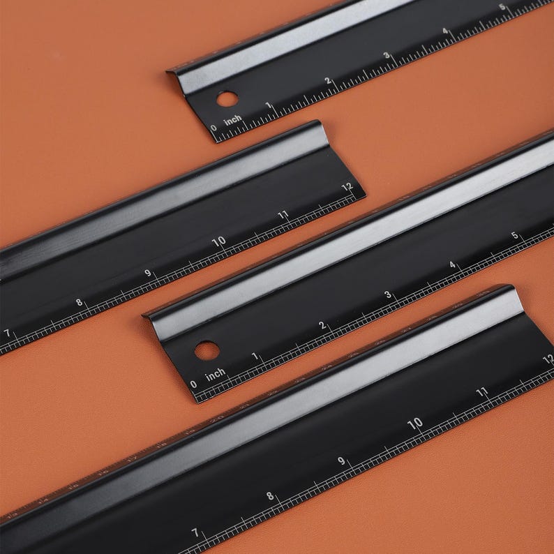 May include: Three black metal rulers with inch markings. The rulers are 12 inches long and have a silver edge.