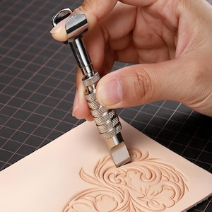 May include: A silver-colored metal swivel knife with a textured, knurled grip handle and a flat, angled blade tip, used for carving intricate, flowing floral patterns into a piece of light beige, smooth-surfaced vegetable-tanned leather.