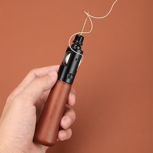 May include: A close-up of a leather sewing awl with a wooden handle and a black metal needle. The awl is being held by a hand and has a white thread attached to the needle.