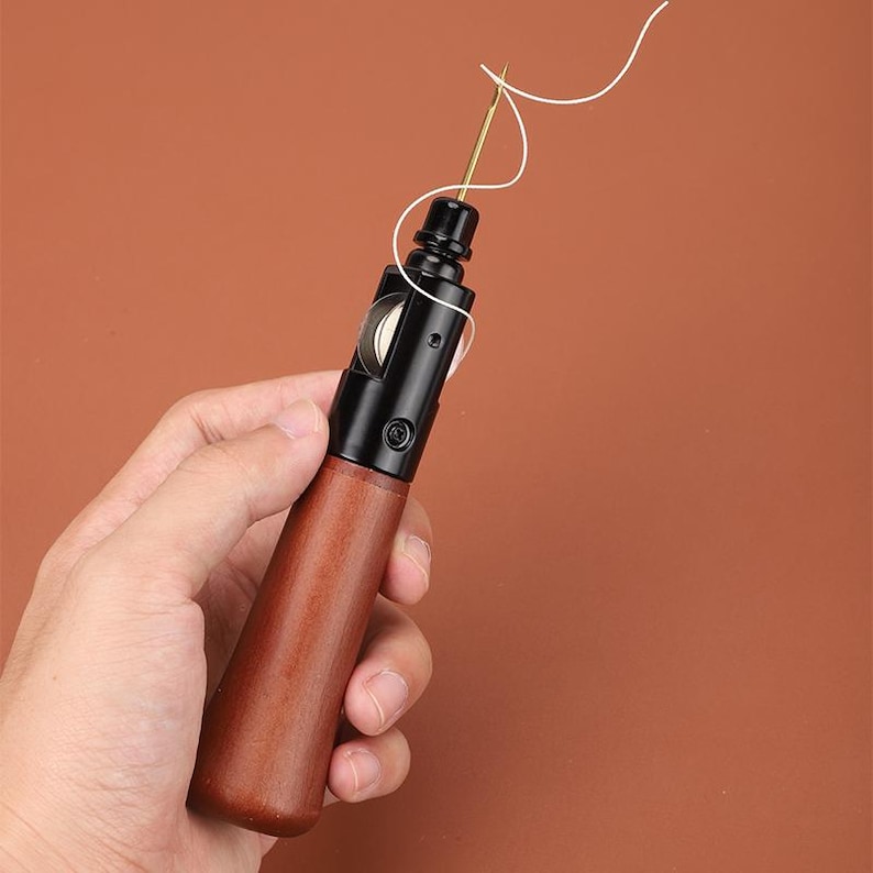 May include: A close-up of a leather sewing awl with a wooden handle and a black metal needle. The awl is being held by a hand and has a white thread attached to the needle.