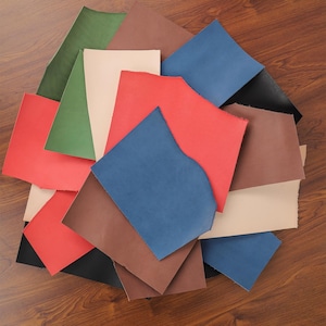 May include: A collection of irregularly shaped leather pieces in various colors, including red, blue, brown, green, and black. The pieces are arranged in a scattered pile, with the edges of the leather showing a raw, unfinished texture.