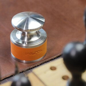May include: A stainless steel tamper with an orange leather band. The band is embossed with the word "ALTA". The tamper has a circular top and base, and is designed for pressing coffee grounds.