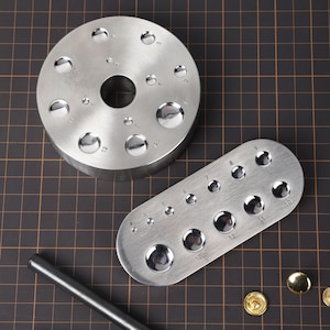 May include: Two metal tools for punching holes in leather. The larger tool is a round disc with a hole in the center and 12 different sized punch holes around the edge. The smaller tool is an oval shape with 10 different sized punch holes. Both tools are made of silver metal.