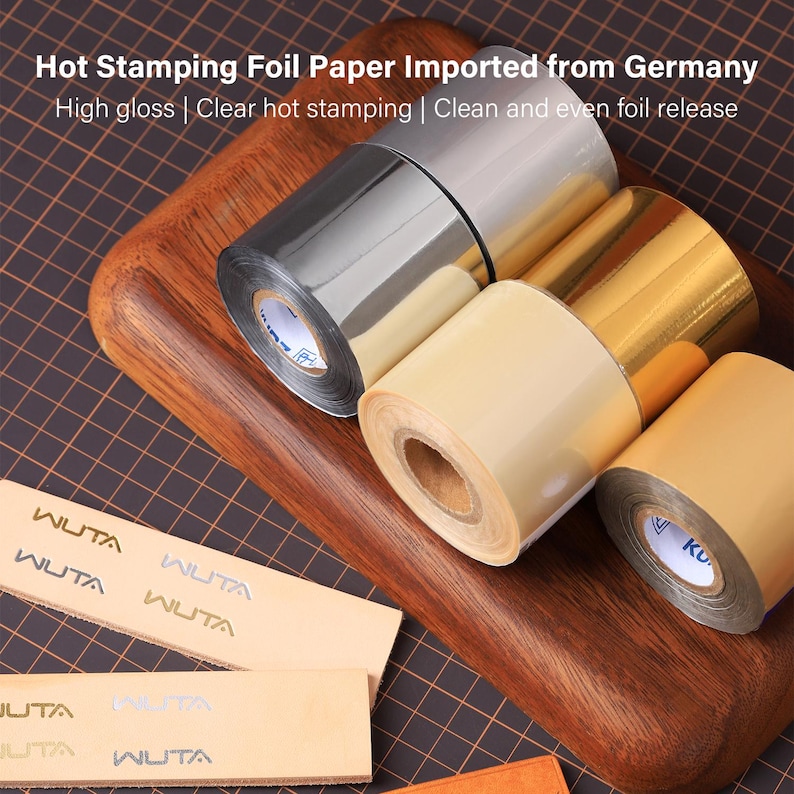 May include: Rolls of hot stamping foil paper in silver, gold, and cream colors are displayed on a wooden cutting board. The text "Hot Stamping Foil Paper Imported from Germany" is visible. Several strips of paper with the word "WUTA" are also present.