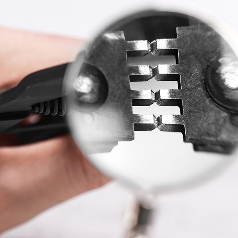 May include: Close-up of a metal wire stripper tool. The tool is silver and black, with a series of rectangular notches visible. The tool is being viewed through a magnifying glass, with a hand holding the tool in the background.