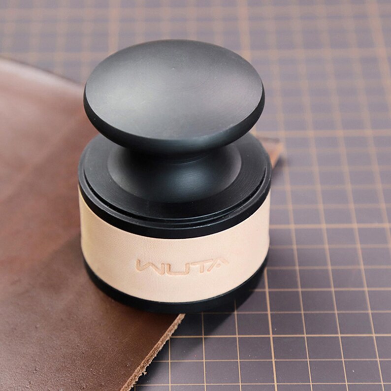 May include: A black leathercraft tool with a rounded top and a light tan leather base. The word "WUTA" is embossed on the leather. The tool is sitting on a piece of brown leather.