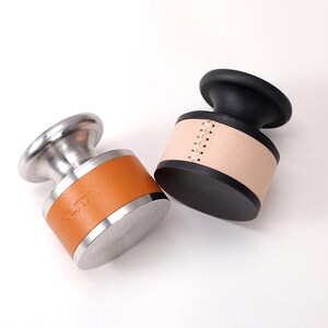 May include: Two bottle stoppers with a modern design. One has a stainless steel base and a tan leather band with the word "WUTA" embossed. The other has a black base and top with a tan leather band and white stitching.