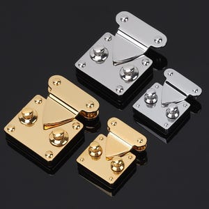 May include: Four metal purse clasps in gold and silver tones. Each clasp features a square base with a central locking mechanism and a hinged top. The clasps are arranged on a reflective black surface.