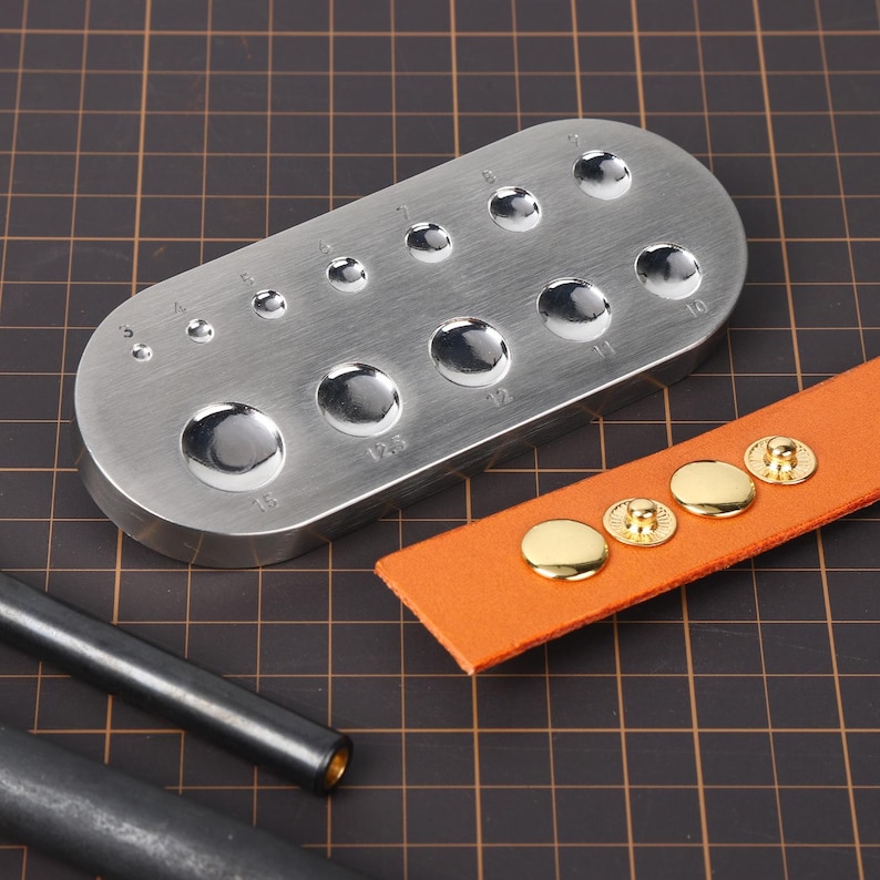 May include: A metal tool with 13 round holes of different sizes for setting leather snaps. The tool is silver and has numbers 3 to 15 printed next to each hole. There are four gold leather snaps on a piece of orange leather.