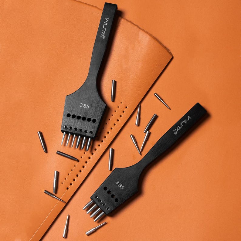 May include: Two black leather hole punch tools with the brand name "MULTA" and the number "3.85" printed on them. The tools are on a brown leather surface with small metal pins scattered around them.