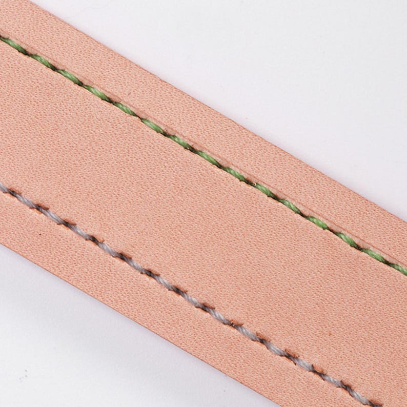 May include: Close-up of a section of light brown leather with two parallel lines of stitching. One line is stitched with light green thread, and the other with light grey thread. The leather has a slightly textured surface.