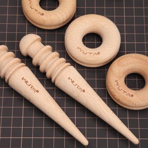 May include: A collection of light-colored wooden leatherworking tools and donut-shaped objects. Two awls and three round tools are visible, all bearing the "WUTA®" brand. The tools are arranged on a black grid surface.