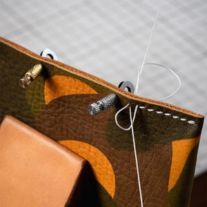 May include: Close-up of a leather craft project with a white thread being sewn through holes in the leather. The leather is brown with a camouflage pattern and has three metal rivets.
