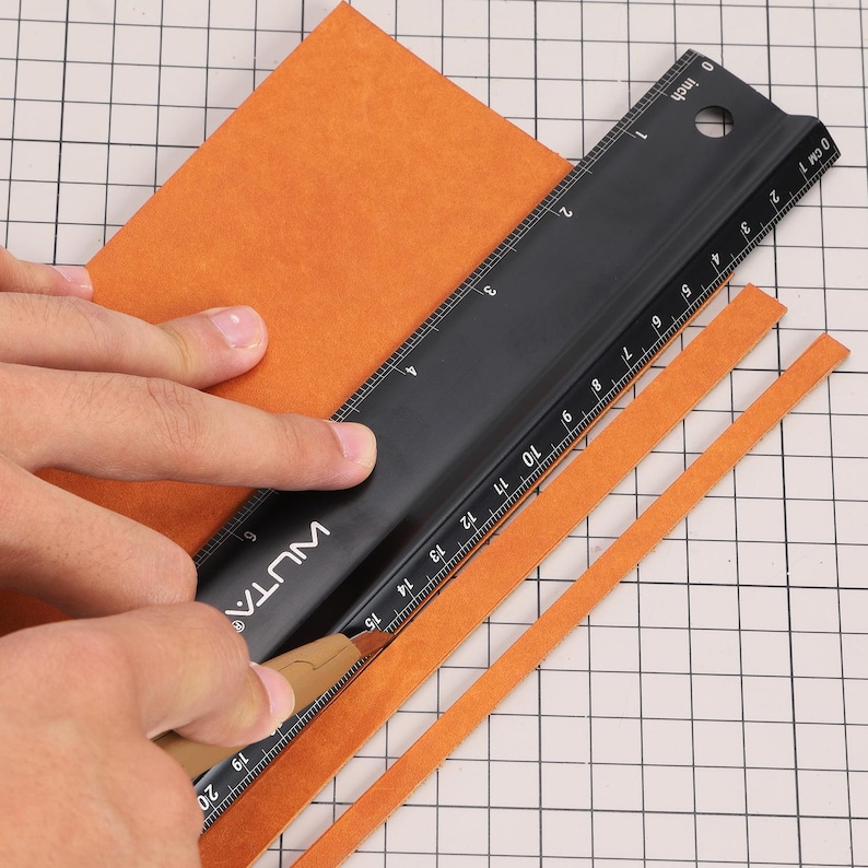 May include: A person using a black ruler and a craft knife to cut strips of orange leather. The ruler is marked in inches and the leather is on a gridded surface.