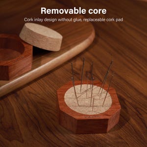 Puede incluir: Un portaagujas de madera con forma hexagonal y una incrustaci&oacute;n de corcho. El soporte contiene varias agujas. El texto "Removable core" y "Cork inlay design without glue, replaceable cork pad" son visibles.
