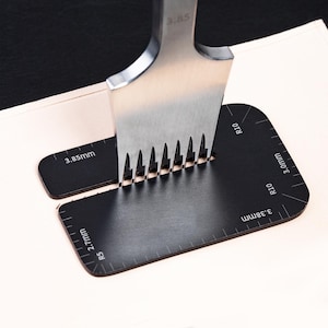 May include: A black leather crafting tool with markings for 2.7mm, 3.0mm, 3.8mm, and 3.85mm. A silver stitching tool with a row of prongs is being used to punch holes in the leather.