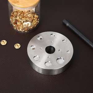 May include: A silver metal tool with a circular base and multiple round holes of different sizes. The tool is used for punching leather or fabric for attaching snaps or other fasteners. The tool has markings indicating the size of each hole in millimeters.
