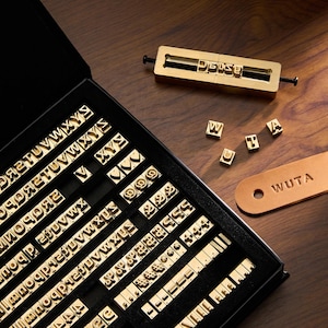 May include: A brass letter stamp set with a variety of capital letters, numbers, and symbols. The set includes a holder with the word "Daisy" stamped, individual letters, and a leather tag with "WUTA" embossed. The set is displayed on a wooden surface.