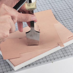 Puede incluir: Una persona usando una herramienta de costura de cuero para perforar agujeros en un trozo de cuero. La herramienta tiene un mango y una cabeza de metal con m&uacute;ltiples p&uacute;as. El cuero se coloca sobre una estera de espuma blanca con un patr&oacute;n de cuadr&iacute;cula.