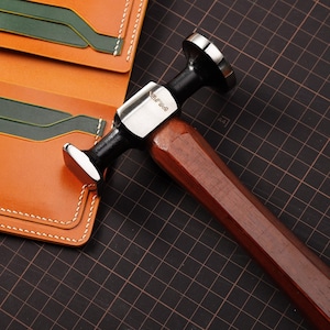 May include: A leathercraft hammer with a dark wooden handle and a black and silver head, next to a tan leather wallet with green card slots. The hammer has the text "VLJM" on the side.
