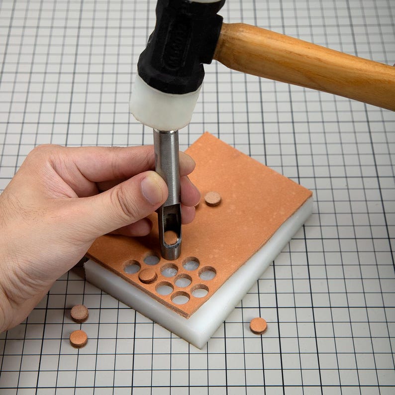 May include: A person using a leather punch tool to create holes in a piece of leather. The tool is being hammered with a mallet. There are several leather circles around the leather piece.